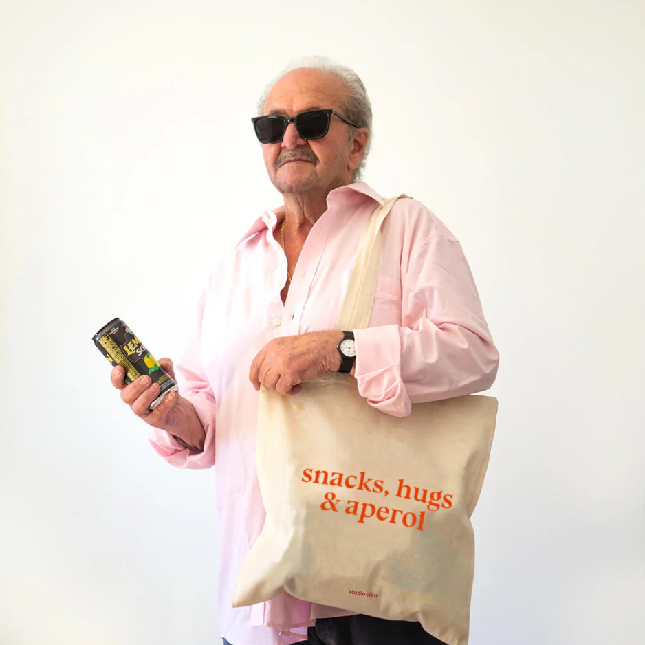 Naturfarbener Jutebeutel mit orangenem Schriftzug "snacks, hugs and aperol" und langen Henkeln, getragen von einem Mann, Design von Studio Ciao
